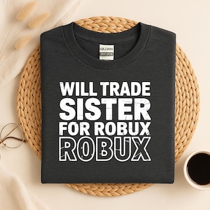 May include: Dark gray t-shirt with the text "WILL TRADE SISTER FOR ROBUX" in white, a humorous statement. The shirt is made of heavy cotton and is displayed on a woven mat, with a cup of coffee and glasses in the background.
