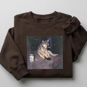 K&ouml;nnte beinhalten: Ein braunes Sweatshirt mit einem aufgedruckten Bild eines Wolfs, der eine Zigarette raucht, mit einer Bierdose daneben. Der Wolf ist in entspannter Pose vor einem dunklen, sternen&uuml;bers&auml;ten Hintergrund dargestellt. Das Sweatshirt ist aus weichem Material.