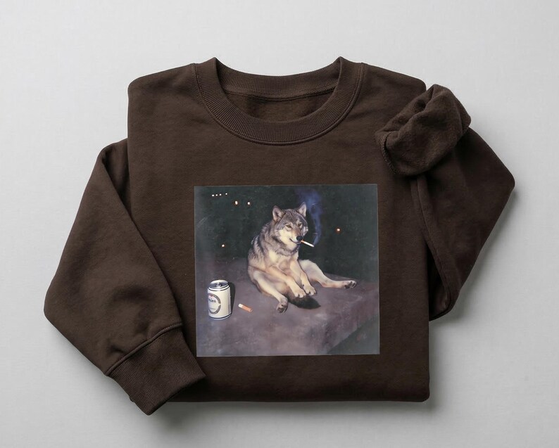 K&ouml;nnte beinhalten: Ein braunes Sweatshirt mit einem aufgedruckten Bild eines Wolfs, der eine Zigarette raucht, mit einer Bierdose daneben. Der Wolf ist in entspannter Pose vor einem dunklen, sternen&uuml;bers&auml;ten Hintergrund dargestellt. Das Sweatshirt ist aus weichem Material.
