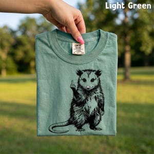 May include: Light green t-shirt with a black graphic of an opossum giving the middle finger. The shirt is held up by a hand. The text "Light Green" is in the top right corner. The shirt has a Comfort Colors tag.