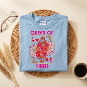Op de afbeelding: Lichtblauw T-shirt met de tekst "QUEEN OF FARTS" en een speelkaartontwerp met een koningin met een kroon. De afbeelding is roze, oranje en rood. Het shirt ligt op een geweven mat.