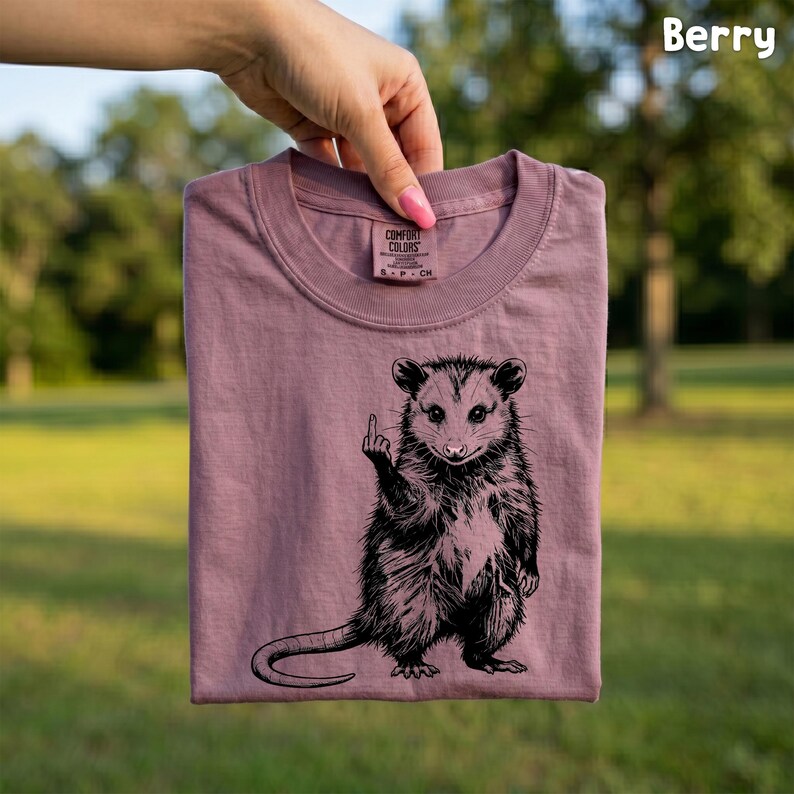 May include: Berry-colored t-shirt featuring a black and white opossum graphic giving the middle finger. The shirt is a crew neck and made of a soft material. The text "Comfort Colors" is visible on the tag.