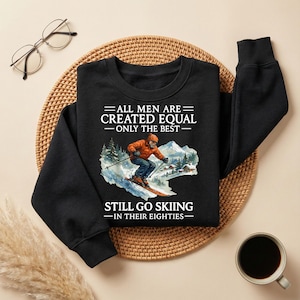 May include: Black sweatshirt with the text "ALL MEN ARE CREATED EQUAL - ONLY THE BEST - STILL GO SKIING - IN THEIR EIGHTIES" and a graphic of a skier. The sweatshirt is on a woven mat with a cup of coffee and glasses.