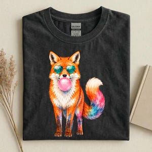 Peut inclure: Un t-shirt à col rond gris anthracite avec un motif de renard orange portant des lunettes de soleil en forme de cœur et faisant une bulle de chewing-gum rose. Le renard a une queue multicolore aux teintes bleues et roses. Étiquette Gildan.