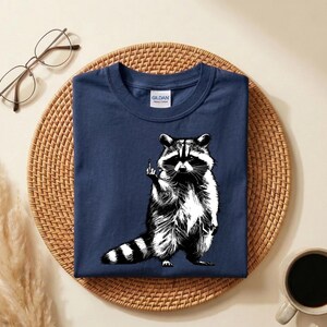 May include: Navy blue t-shirt featuring a black and white illustration of a raccoon giving the middle finger. The raccoon has a striped tail and is standing upright. The shirt has a crew neck and a Gildan tag.