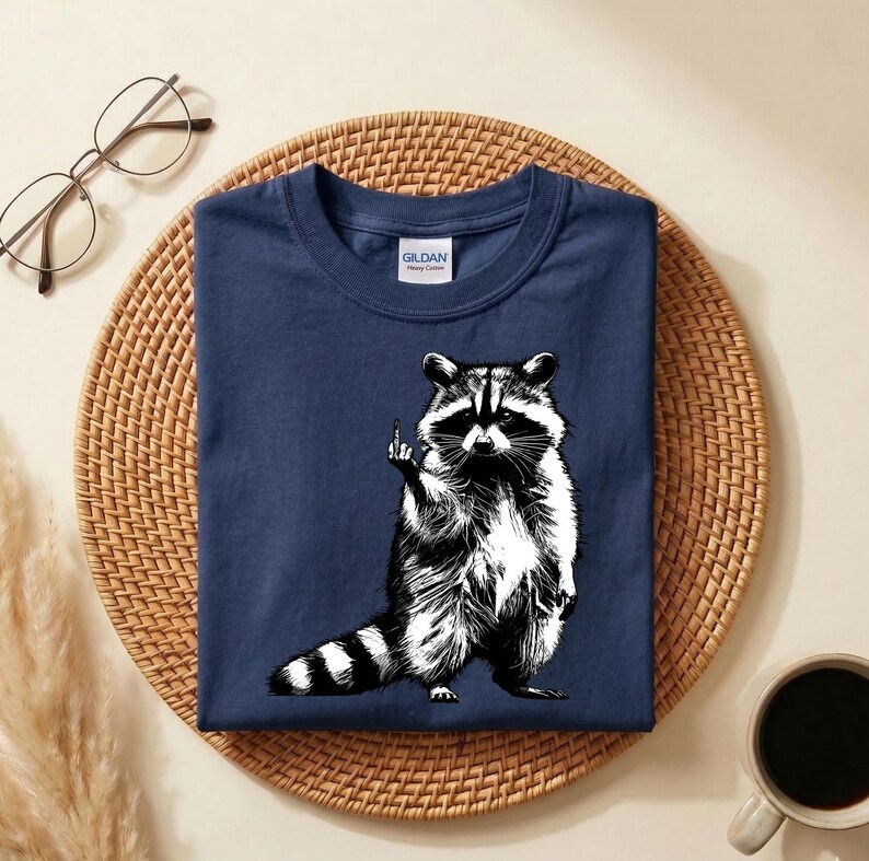 May include: Navy blue t-shirt featuring a black and white illustration of a raccoon giving the middle finger. The raccoon has a striped tail and is standing upright. The shirt has a crew neck and a Gildan tag.
