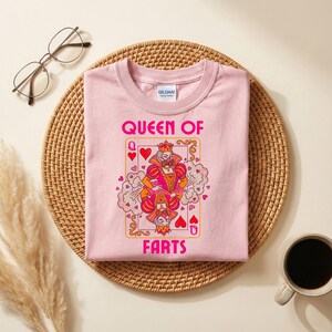 Op de afbeelding: Een lichtroze t-shirt met een speelkaartontwerp met een koningin met een kroon. De tekst "QUEEN OF FARTS" is roze gedrukt. Het shirt ligt op een geweven mat met een bril en een koffiekopje.