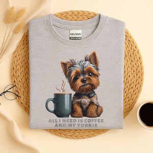May include: A light gray t-shirt featuring a cartoon Yorkshire Terrier with a coffee mug. The dog is brown, black, and tan, with a small black bow tie. The text "ALL I NEED IS COFFEE AND MY YORKIE" is printed below the image.