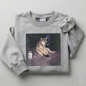 K&ouml;nnte beinhalten: Ein hellgraues Sweatshirt mit einem Grafikdruck eines Wolfs, der eine Zigarette raucht, mit einer Bierdose und einer Zigarette. Der Wolf ist in einer entspannten Pose dargestellt. Das Sweatshirt ist aus weichem Material.