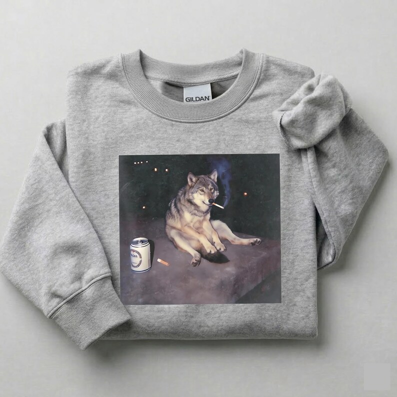 K&ouml;nnte beinhalten: Ein hellgraues Sweatshirt mit einem Grafikdruck eines Wolfs, der eine Zigarette raucht, mit einer Bierdose und einer Zigarette. Der Wolf ist in einer entspannten Pose dargestellt. Das Sweatshirt ist aus weichem Material.
