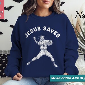 May include: Navy blue sweatshirt with the text "JESUS SAVES" above an illustration of Jesus in a baseball uniform throwing a ball. The sweatshirt has a crew neck and long sleeves. The words "MORE COLOR AND STYLE OPTIONS" are at the bottom.