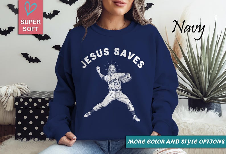 May include: Navy blue sweatshirt with the text "JESUS SAVES" above an illustration of Jesus in a baseball uniform throwing a ball. The sweatshirt has a crew neck and long sleeves. The words "MORE COLOR AND STYLE OPTIONS" are at the bottom.