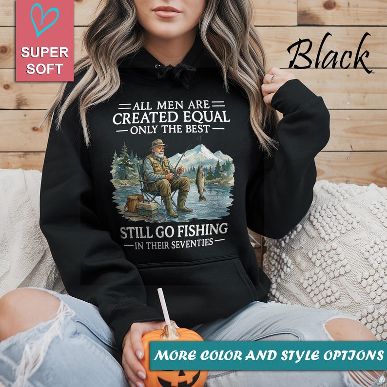 Op de afbeelding: Zwarte hoodie met een afbeelding van een visser, bergen en de tekst "ALL MEN ARE CREATED EQUAL - ONLY THE BEST - STILL GO FISHING - IN THEIR SEVENTIES." Een roze hart en de woorden "SUPER SOFT" staan linksboven.