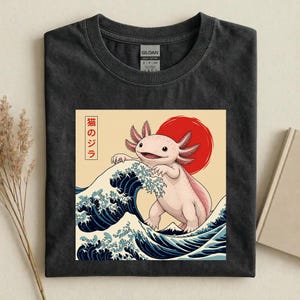 May include: A charcoal gray t-shirt featuring a graphic print of a light pink axolotl standing on a large blue ocean wave. Behind the axolotl is a large red circle. The design includes vertical Japanese text in a white box on the left side.