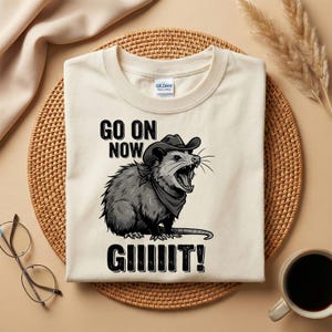 Puede incluir: Camiseta color crema con un gráfico de una zarigüeya con sombrero de vaquero y pañuelo, con el texto "GO ON NOW GIIIIIT!". La zarigüeya está representada en negro y gris, con la boca abierta gritando.