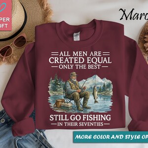 Op de afbeelding: Bordeaux sweatshirt met een afbeelding van een visser die op een stoel zit te vissen. De tekst luidt: "ALLE MANNEN ZIJN GELIJK GESCHAPEN - ALLEEN DE BESTEN - GAAN NOG VISSEN - IN HUN ZEVENTIG." De sweatshirt heeft een ronde hals.