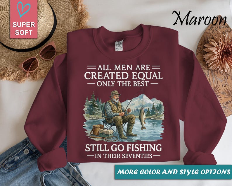 Op de afbeelding: Bordeaux sweatshirt met een afbeelding van een visser die op een stoel zit te vissen. De tekst luidt: "ALLE MANNEN ZIJN GELIJK GESCHAPEN - ALLEEN DE BESTEN - GAAN NOG VISSEN - IN HUN ZEVENTIG." De sweatshirt heeft een ronde hals.