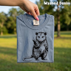 May include: A washed denim blue t-shirt featuring a black and white illustration of an opossum giving the middle finger. The shirt is a Comfort Colors brand and is being held up by a hand. The text "Washed Denim" is at the top.