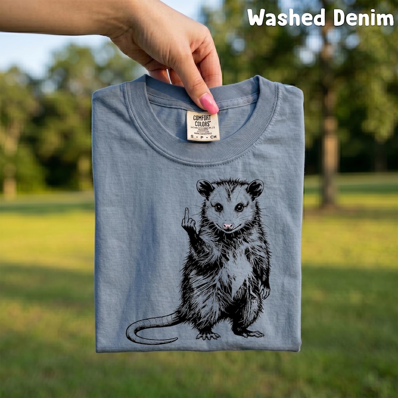 May include: A washed denim blue t-shirt featuring a black and white illustration of an opossum giving the middle finger. The shirt is a Comfort Colors brand and is being held up by a hand. The text "Washed Denim" is at the top.