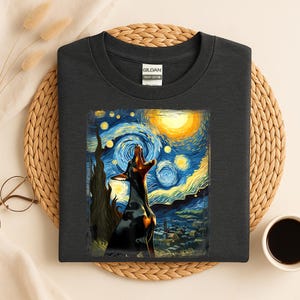 Puede incluir: Camiseta gris oscuro con un Doberman Pinscher aullando a la luna, sobre un cielo nocturno arremolinado que recuerda a "La noche estrellada" de Van Gogh. El dise&ntilde;o incluye un sol amarillo y una escena de pueblo.