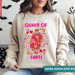 Op de afbeelding: Zandkleurige sweatshirt met een speelse speelkaartafbeelding. De kaart toont een cartoon koningin met de tekst "QUEEN OF FARTS" in roze. De sweatshirt heeft lange mouwen en een ronde hals.