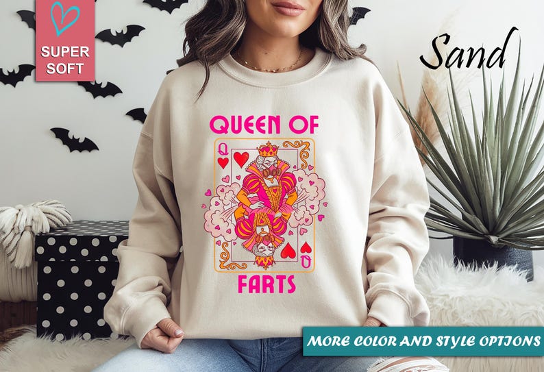 Op de afbeelding: Zandkleurige sweatshirt met een speelse speelkaartafbeelding. De kaart toont een cartoon koningin met de tekst "QUEEN OF FARTS" in roze. De sweatshirt heeft lange mouwen en een ronde hals.