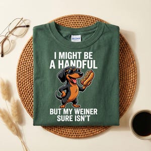 Op de afbeelding: Donkergroen t-shirt met een cartoon teckel met een zonnebril en een hotdog. De tekst luidt "I MIGHT BE A HANDFUL BUT MY WEINER SURE ISN'T".