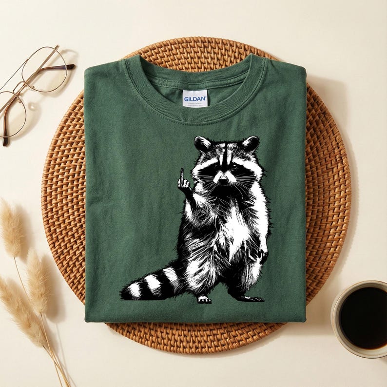 May include: A dark green t-shirt featuring a black and white illustration of a raccoon giving the middle finger. The raccoon is standing upright with its arms at its sides. The shirt is folded on a woven mat.