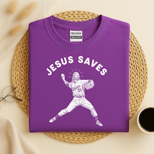 May include: A purple t-shirt with the words "JESUS SAVES" in white arched across the top. Below the text is a graphic of Jesus as a baseball pitcher. He is wearing a baseball uniform with the word "Heaven" and the number "01".