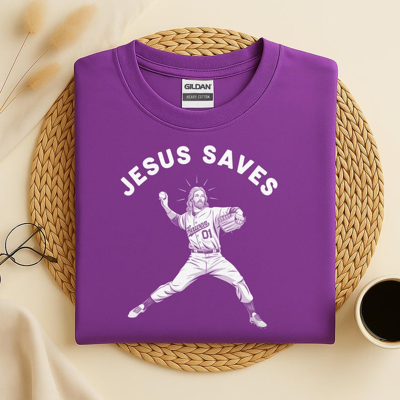 May include: A purple t-shirt with the words "JESUS SAVES" in white arched across the top. Below the text is a graphic of Jesus as a baseball pitcher. He is wearing a baseball uniform with the word "Heaven" and the number "01".