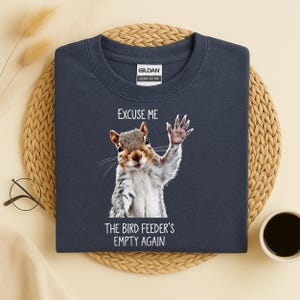 Op de afbeelding: Marineblauwe sweatshirt met een afbeelding van een wuivend eekhoorntje en de tekst "EXCUSE ME THE BIRD FEEDER'S EMPTY AGAIN." De sweatshirt is van het merk Gildan Heavy Cotton.