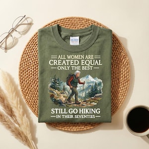 Könnte beinhalten: Olivgrünes T-Shirt mit dem Text "ALL WOMEN ARE CREATED EQUAL - ONLY THE BEST - STILL GO HIKING - IN THEIR SEVENTIES." Es zeigt eine Grafik einer Frau, die in den Bergen wandert. Präsentiert auf einer gewebten Matte.