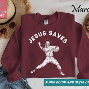 May include: Maroon sweatshirt with the text "JESUS SAVES" in white. A graphic of Jesus pitching a baseball is below the text. The sweatshirt has the words "MORE COLOR AND STYLE OPTIONS" at the bottom.