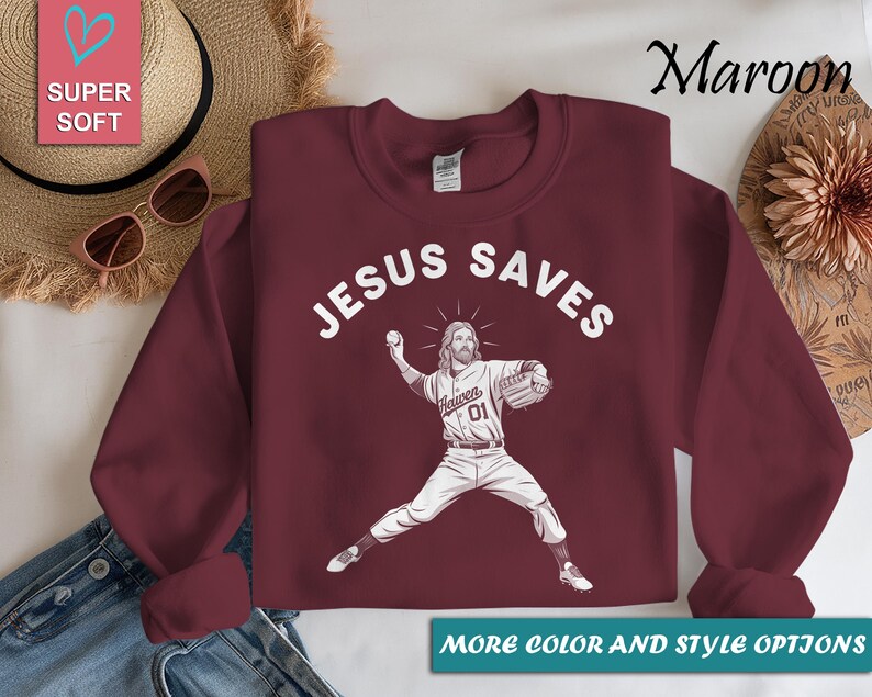 May include: Maroon sweatshirt with the text "JESUS SAVES" in white. A graphic of Jesus pitching a baseball is below the text. The sweatshirt has the words "MORE COLOR AND STYLE OPTIONS" at the bottom.