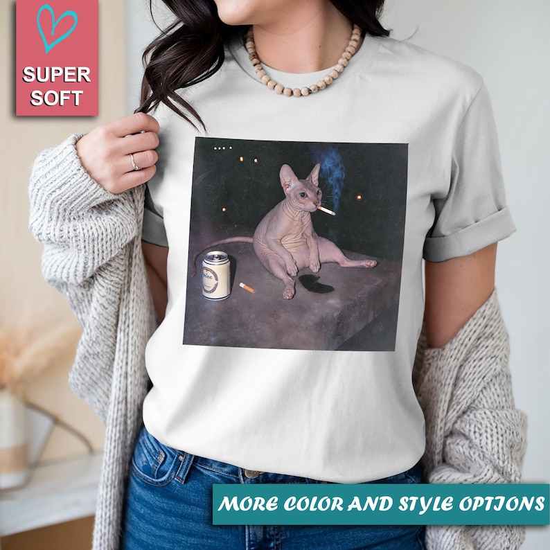 May include: A white short-sleeve t-shirt featuring a graphic print of a hairless Sphynx cat sitting on a dark surface. The cat has a cigarette in its mouth, and a beverage can and another cigarette are placed on the surface next to it.