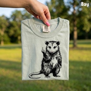 May include: A light green Comfort Colors t-shirt featuring a black and white opossum graphic giving the middle finger. The opossum is standing upright with its tail curled. The shirt has a crew neck and short sleeves.