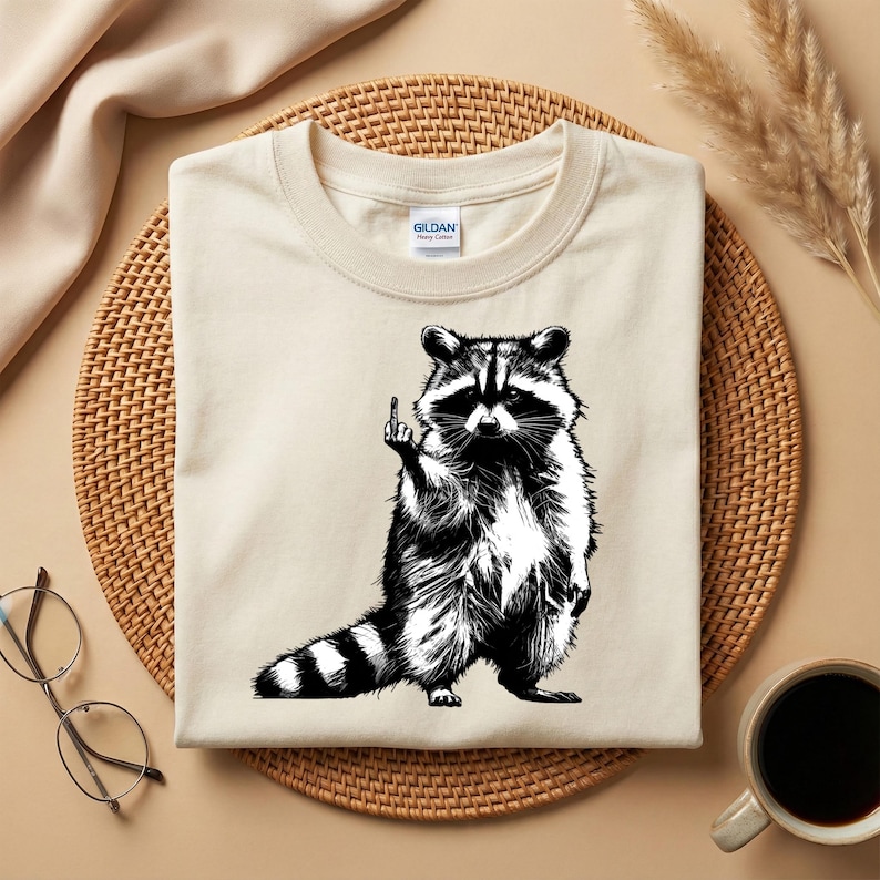 May include: A cream-colored t-shirt featuring a black and white illustration of a raccoon giving the middle finger. The raccoon is standing upright with its tail striped. The shirt has a crew neck and a small tag.