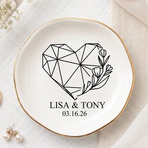 May include: A white ceramic ring dish with a gold rim, featuring a geometric heart design and floral accents in black. The dish is personalized with the names "LISA & TONY" and the date "03.16.26".