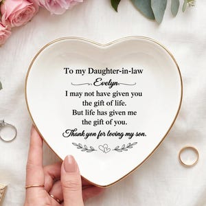 Peut inclure: Un vide-poche en céramique blanche en forme de cœur avec un bord doré, portant l'inscription noire suivante : To my Daughter-in-law Evelyn, I may not have given you the gift of life. But life has given me the gift of you. Thank you for loving my son.
