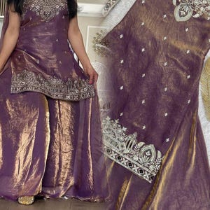 May include: A purple and gold metallic sleeveless tunic and wide-leg trousers set. The tunic features intricate silver and white embroidery around the neckline and hem, with scattered floral motifs. The fabric has a shimmering, textured finish.