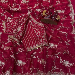 May include: A deep red saree with intricate gold floral embroidery. The saree features a matching blouse with detailed gold embellishments and a scalloped border. Decorative figurines are placed on the saree.