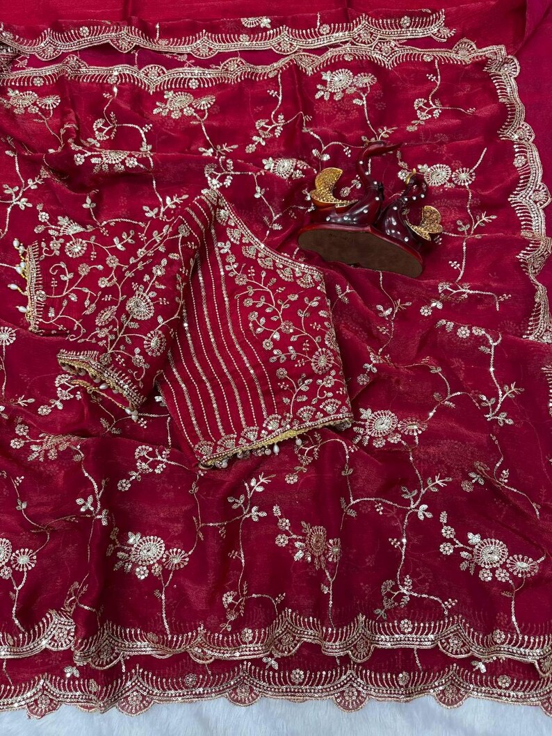 May include: A deep red saree with intricate gold floral embroidery. The saree features a matching blouse with detailed gold embellishments and a scalloped border. Decorative figurines are placed on the saree.
