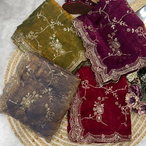 May include: Four colorful, embroidered shawls are displayed on a woven mat. The shawls are in shades of olive green, deep purple, charcoal gray, and burgundy. Each shawl features intricate floral embroidery and a decorative, scalloped border.