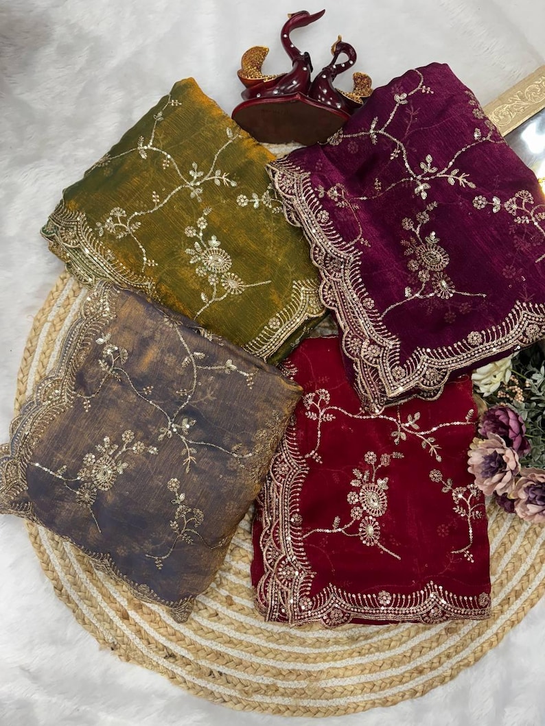 May include: Four colorful, embroidered shawls are displayed on a woven mat. The shawls are in shades of olive green, deep purple, charcoal gray, and burgundy. Each shawl features intricate floral embroidery and a decorative, scalloped border.