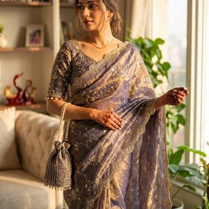 May include: A lavender sari with gold embroidery and a matching blouse. The sari features a scalloped border and is paired with a beaded drawstring bag. The blouse has short sleeves and intricate detailing. The outfit is elegant and suitable for special occasions.