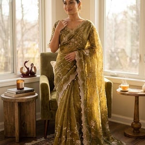 May include: A woman wearing a gold-green sari with intricate embroidery and a scalloped border. The sari is draped elegantly, with a matching blouse. The scene is set indoors, with natural light illuminating the sari's details.