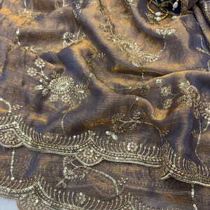 May include: Close-up of a luxurious, draped fabric in shades of brown and gold. The material is embellished with intricate gold embroidery, including floral patterns and delicate sequin detailing. A blue and gold embellished sleeve is visible in the background.