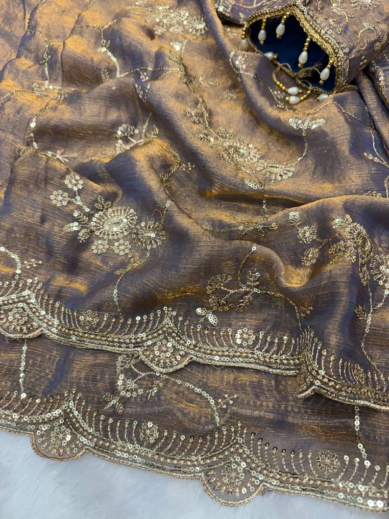 May include: Close-up of a luxurious, draped fabric in shades of brown and gold. The material is embellished with intricate gold embroidery, including floral patterns and delicate sequin detailing. A blue and gold embellished sleeve is visible in the background.