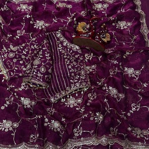 May include: A deep purple saree with intricate gold embroidery. The fabric is adorned with floral and vine patterns, and a matching blouse piece is included. The saree has a scalloped border and is draped with a pair of embellished sandals.