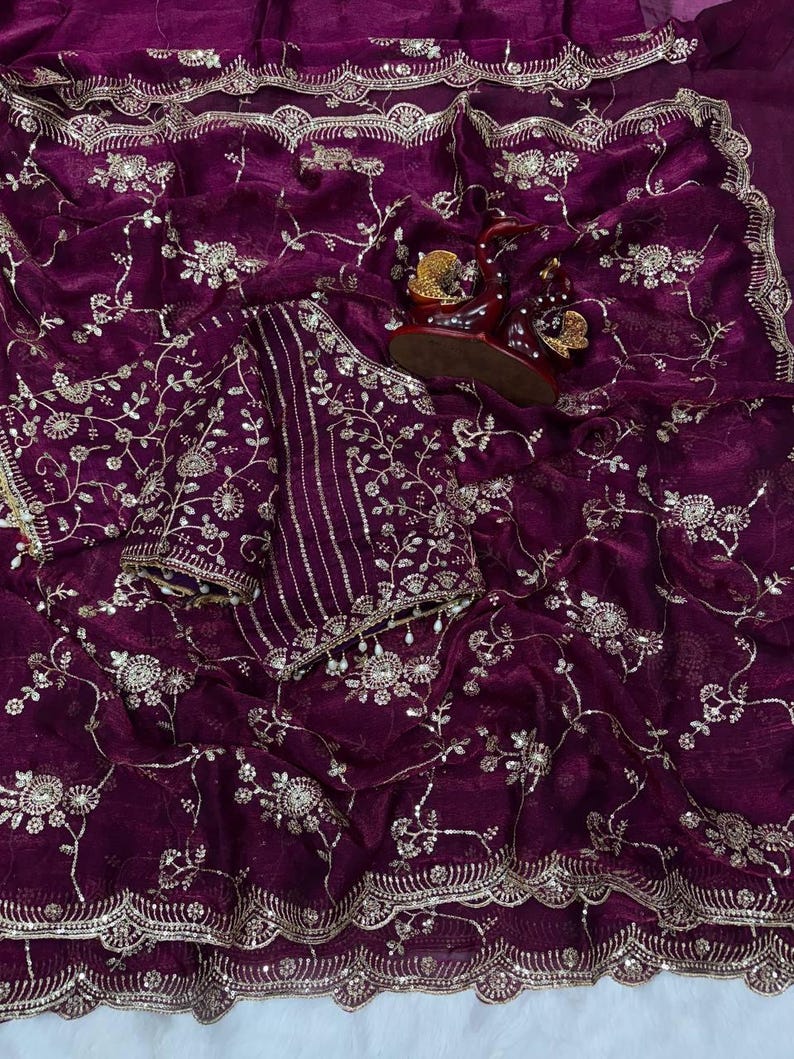 May include: A deep purple saree with intricate gold embroidery. The fabric is adorned with floral and vine patterns, and a matching blouse piece is included. The saree has a scalloped border and is draped with a pair of embellished sandals.